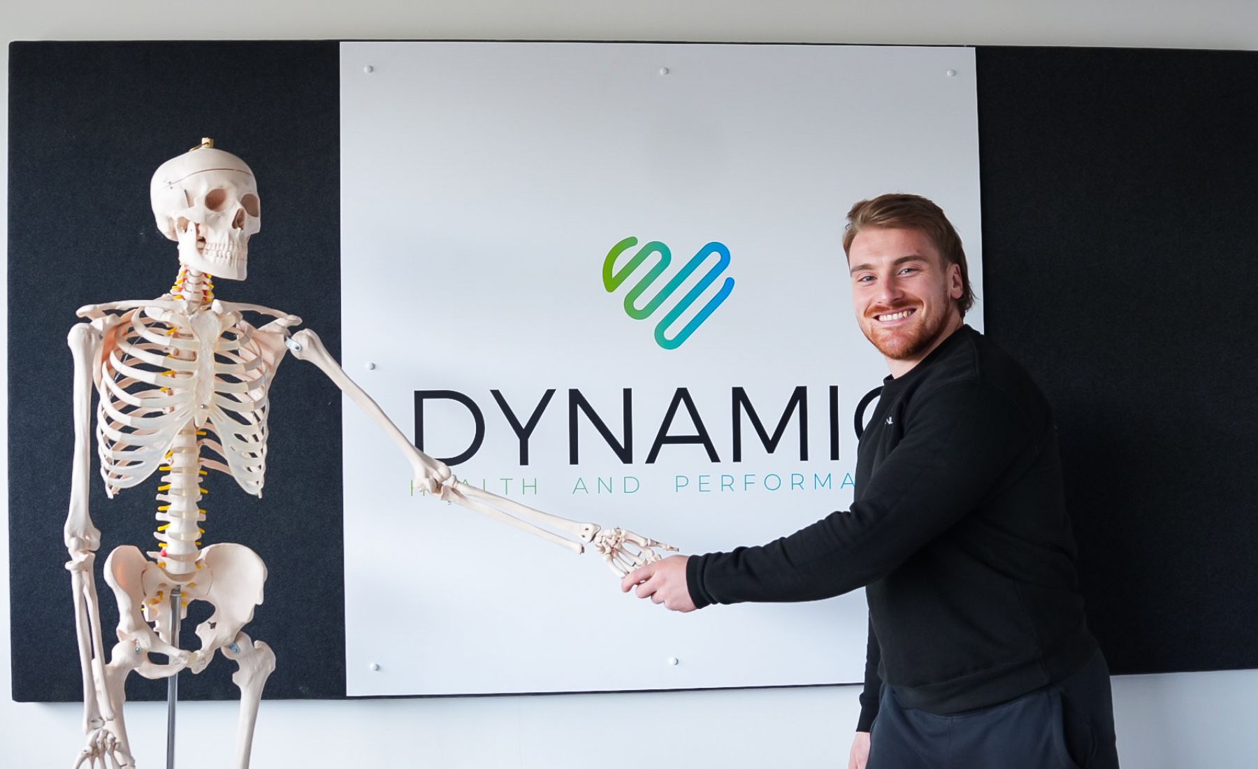 A man smiling and shaking hands with a skeleton in front of a branded sign.