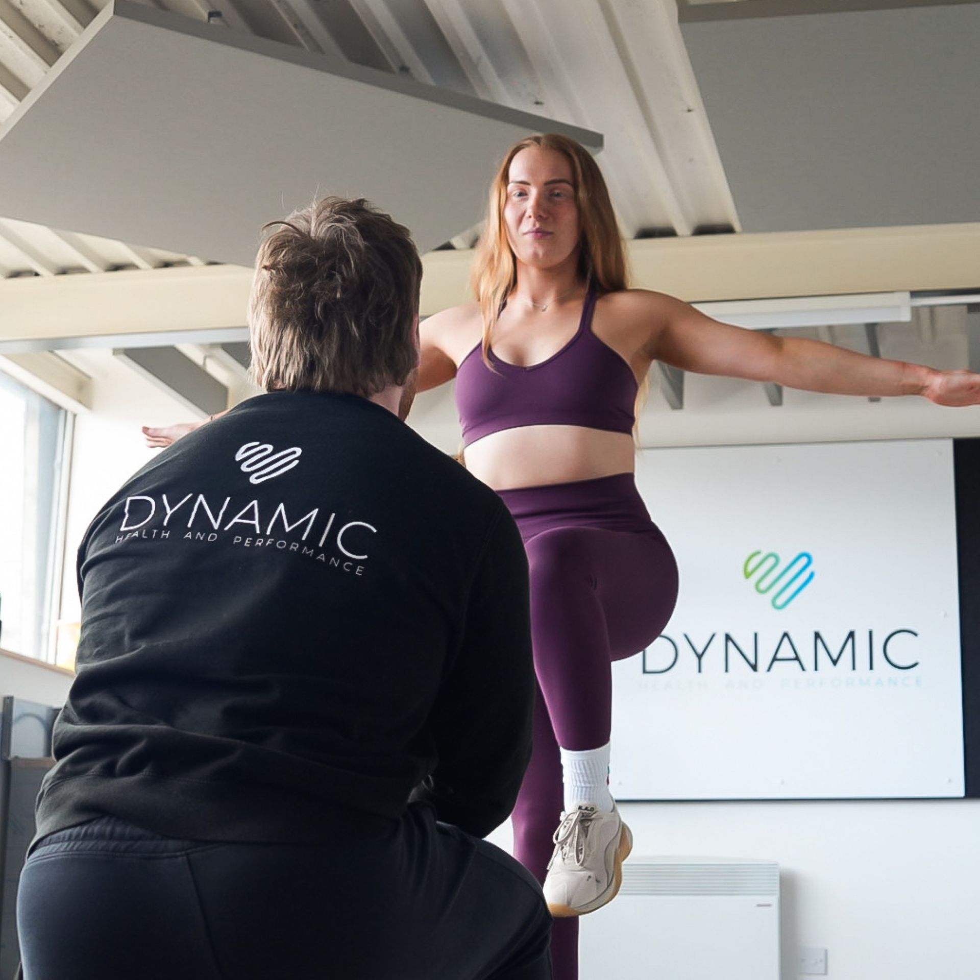 A woman balances on one leg while a trainer observes in a gym setting.