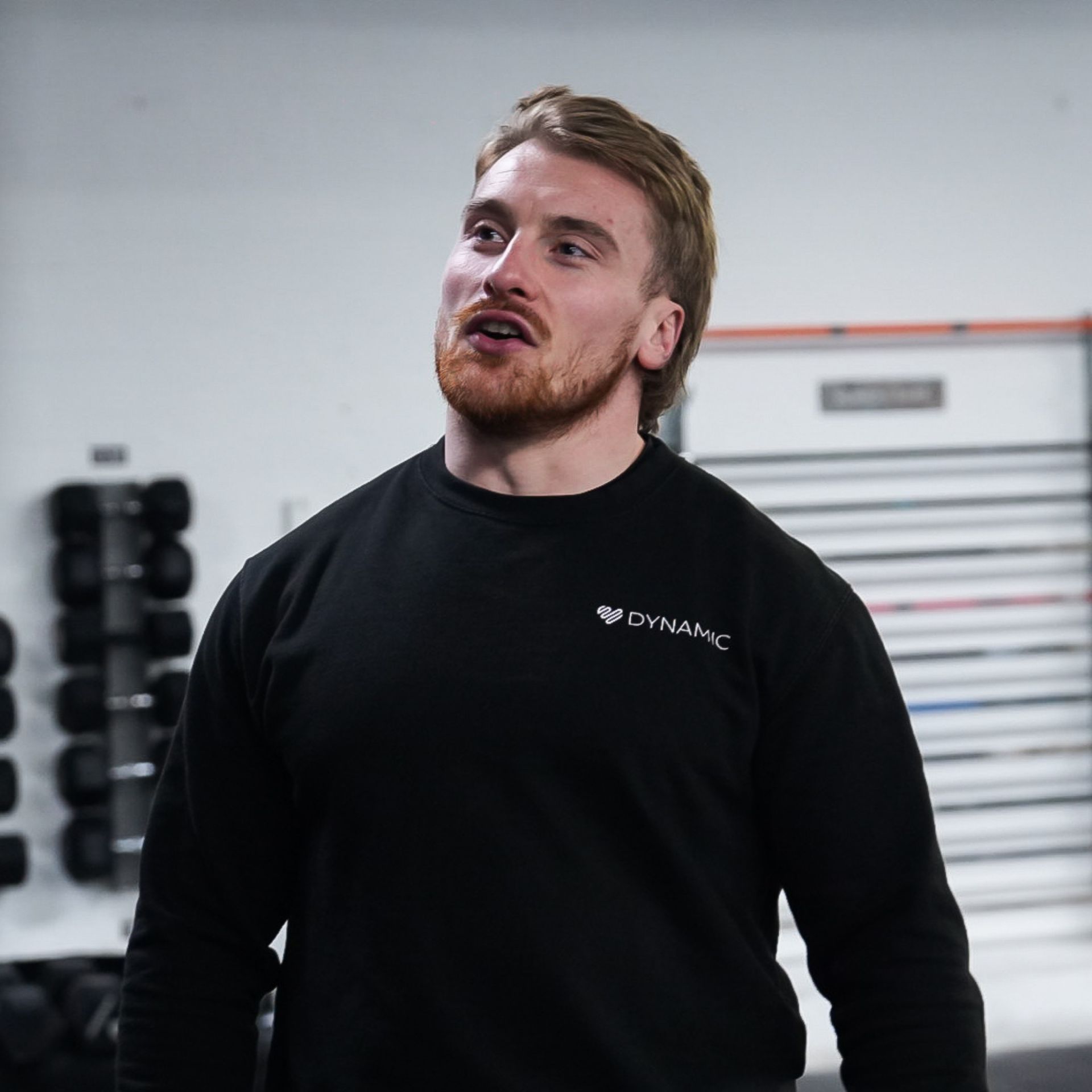Man wearing a black sweater talks in a gym setting with weights in the background.