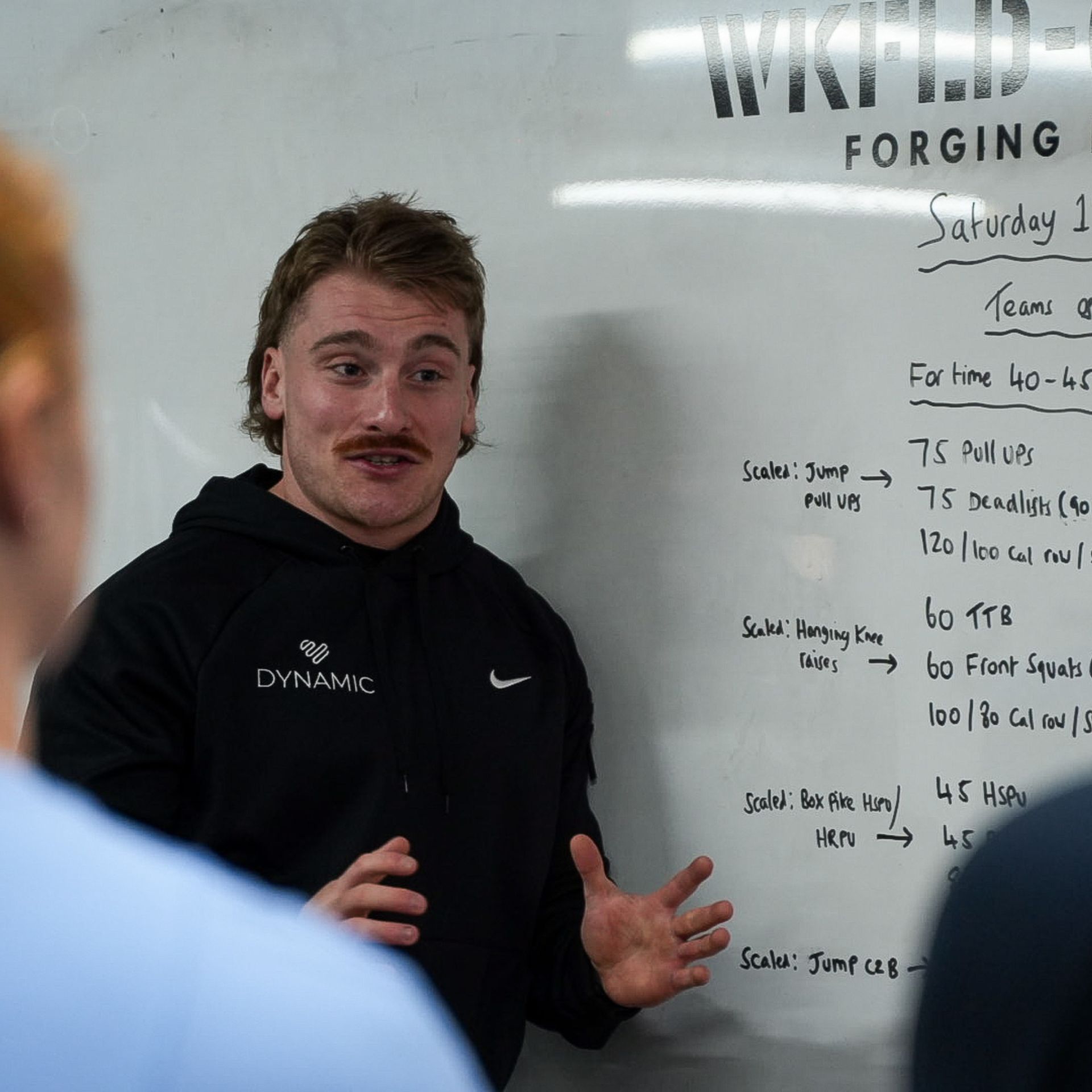 A person in a black hoodie talks and gestures in front of a whiteboard with notes.